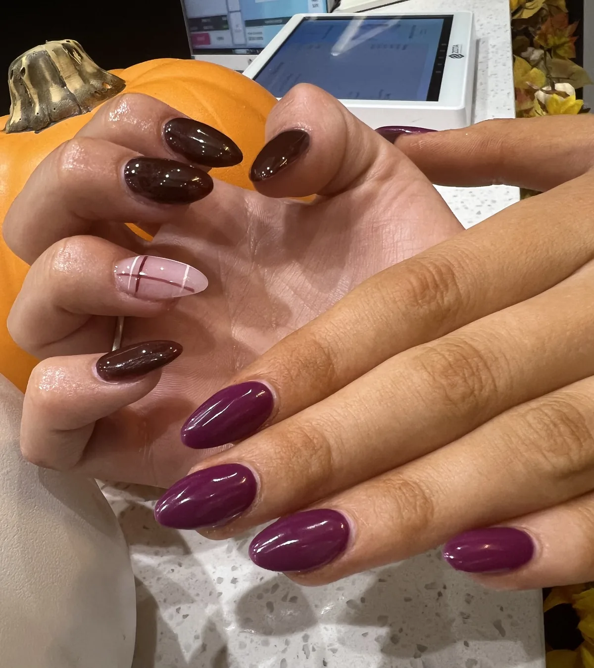 Plum and burgundy almond nails with a pink plaid accent beside a pumpkin at Apple Nailed It Spa
